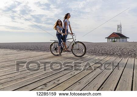 Stock Photo - Two girls riding double on a single bike on a beach boardwalk; Toronto, Ontario, Canada. Fotosearch