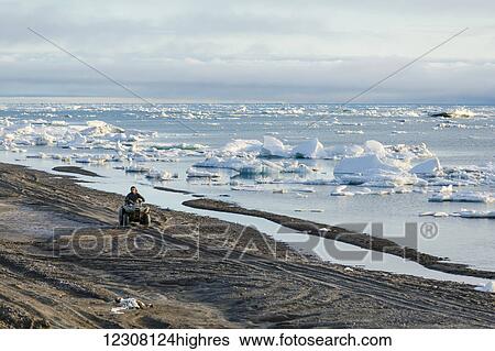 Stock Photograph - Two native Alaskan men drive an ATV down a sand beach along the Arctic Ocean filled with sea ice in summer, 0rth Slope, Arctic Alaska, USA. Fotosearch