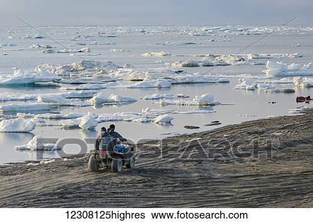 Stock Photograph - Two native Alaskan men drive an ATV down a sand beach along the Arctic Ocean filled with sea ice in summer, 0rth Slope, Arctic Alaska, USA. Fotosearch