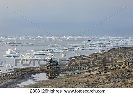 Stock Photograph - Two native Alaskan men drive an ATV down a sand beach along the Arctic Ocean filled with sea ice in summer, 0rth Slope, Arctic Alaska, USA. Fotosearch