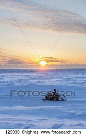 Stock Photograph - Two native Alaskans drive a snow mobile through a stark snow covered landscape at sunset, Barrow, North Slope, Arctic Alaska, Winter. Fotosearch