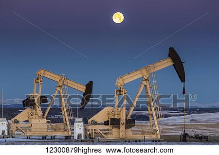 Stock Photograph - Two pump jacks at dusk in a snow covered field with a full moon in a clear sky and snow covered foothills and mountains in the background; Alberta, Canada. Fotosearch
