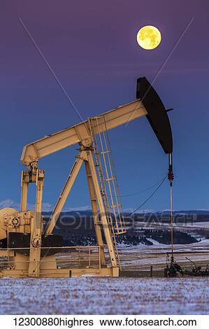 Stock Photograph - Two pump jacks at dusk in a snow covered field with a full moon in a clear sky and snow covered foothills and mountains in the background; Alberta, Canada. Fotosearch
