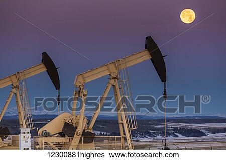 Stock Photograph - Two pump jacks at dusk in a snow covered field with a full moon in a clear sky and snow covered foothills and mountains in the background; Alberta, Canada. Fotosearch