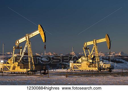 Stock Photograph - Two pump jacks at sunrise reflecting the warm morning light in a snow covered field and snow covered foothills and mountains in the background lit by the morning sun; Alberta, Canada. Fotosearch
