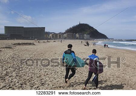 Stock Image - Two surfers in wetsuits walking towards the waves with the two glass cubes of the Kursaal, cultural centre, next to the Rio Urumea and the Zurriola beach in the Gros district; San Sebastian, Spain. Fotosearch
