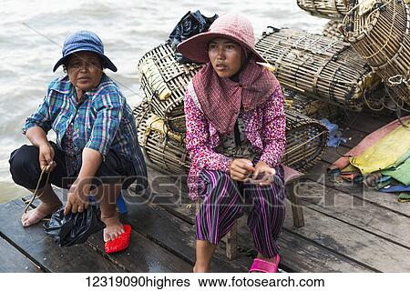 Stock Photograph - Two women sit on a wooden dock at the water's edge with a pile of crab traps behind them, local life in the famous crab market; Kep, Cambodia. Fotosearch