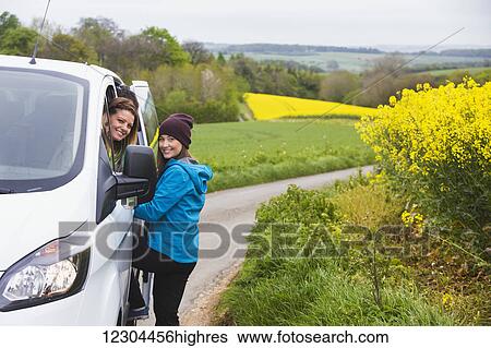 Two young women pose at their vehicle while driving through the English countryside which is covered in vibrant yellow Rapeseed flowers; Cotswolds, England View Large Photo Image Stock Photograph - Two young women pose at their vehicle while driving through the English countryside which is covered in vibrant yellow Rapeseed flowers; Cotswolds, England. Fotosearch