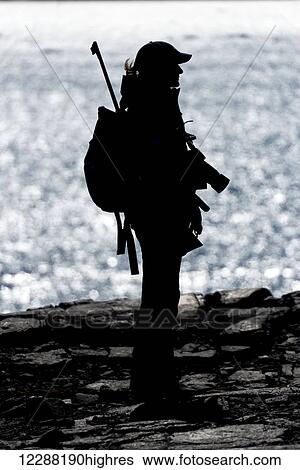 Stock Photograph - Underexposed silhouette of a woman in a peaked cap carrying a backpack, camera and rifle while standing on a rocky cliff, Arctic Ocean with the sun sparkling on it in the background; Spitsbergen, Svalbard, Norway. Fotosearch