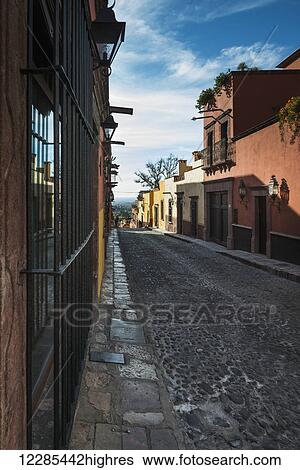 Stock Photograph - View down a narrow cobblestone street with bars in front of windows on buildings; San Miguel de Allende, Guanajuato, Mexico. Fotosearch