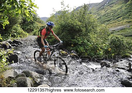 Stock Photograph - Woman rides a full suspension mountain bike across a stream on the Devil's Pass Trail in the Chugach National Forest, Kenai Peninsula, South-central Alaska. Fotosearch