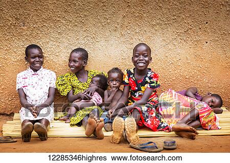 Stock Photograph - Young girls and small children sitting together on a mat against a wall; Gulu, Uganda. Fotosearch