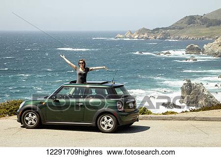 Stock Photograph - Young woman smiling and standing up out of the sunroof of her car on the side of coastal drive Highway one, California coast; California, United States of America. Fotosearch