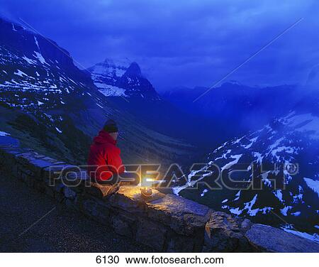 Stock Image - Hiker with lantern sitting on rock wall over mountain valley view. Fotosearch