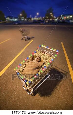 Stock Image - Parking lot with Baby Jesus in shopping cart decorated with lights depicting the loss of the true meaning of Christmas. Fotosearch