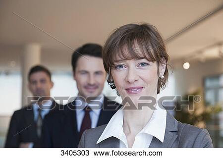 Stock Image - Business woman smiling at camera, two business men behind . Fotosearch