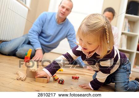 Stock Photo - Girl playing with toys with her parents behind her . Fotosearch