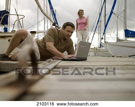 Stock Photograph - Man using laptop at harbor with woman walking in background . Fotosearch