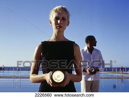 Stock Image - Portrait of woman holding flashlight with man in background . Fotosearch