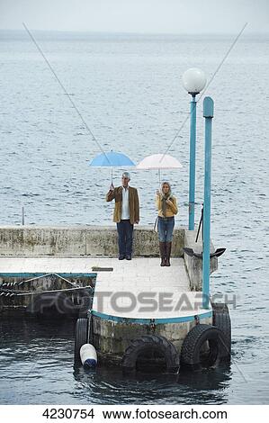 Senior couple standing with umbrella on a pier, Italy, Gardone Riviera View Large Photo Image Picture - Senior couple standing with umbrella on a pier, Italy, Gardone Riviera . Fotosearch