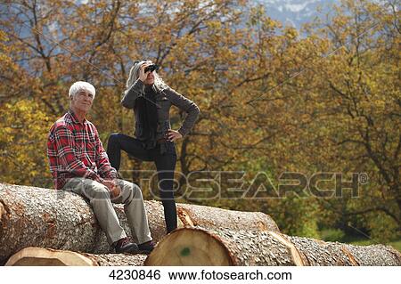 Stock Photograph - Senior couple with binoculars on tree logs, Italy, Telve, Dolomites . Fotosearch