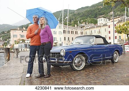 Stock Image - Senior couple with vintage car and umbrella at harbor, Italy, Gargnano, Lake Garda . Fotosearch