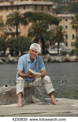 Stock Photography - Senior man sitting by the water holding book, Italy, Rapallo . Fotosearch