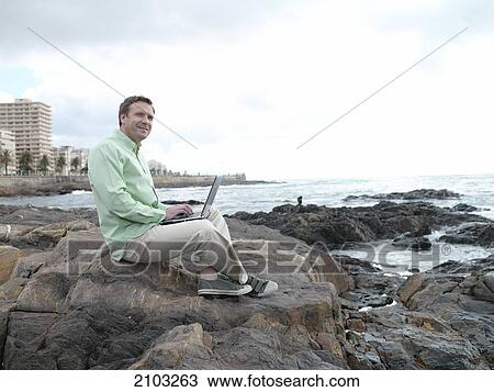 Stock Image - Side profile of man using laptop at coast . Fotosearch