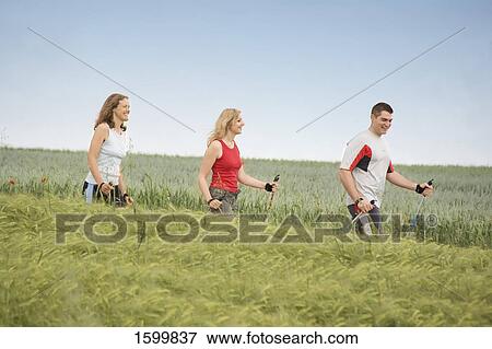 Stock Photo - Side profile of two women and man walking with hiking poles in field . Fotosearch