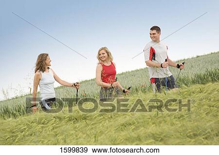 Stock Photo - Side profile of two women and man walking with hiking poles in field . Fotosearch
