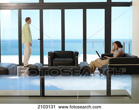 Stock Image - Side profile of woman using laptop on couch with man standing by window . Fotosearch