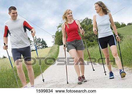 Two women and man walking with hiking poles on dirt road View Large Photo Image Picture - Two women and man walking with hiking poles on dirt road . Fotosearch