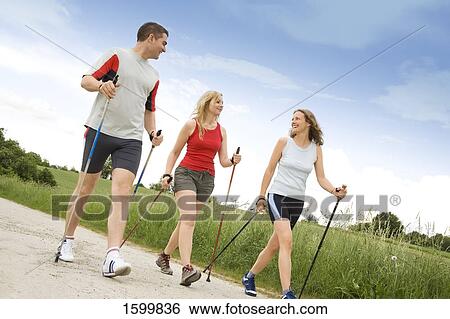 Stock Photograph - Two women and man walking with hiking poles on dirt road . Fotosearch
