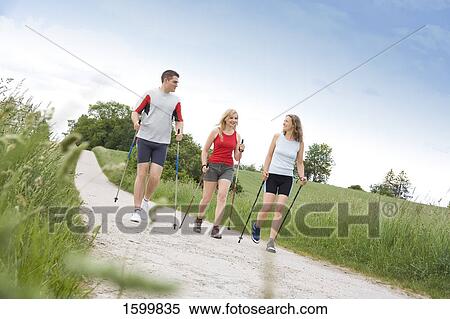 Stock Photography - Two women and man walking with hiking poles on dirt road . Fotosearch
