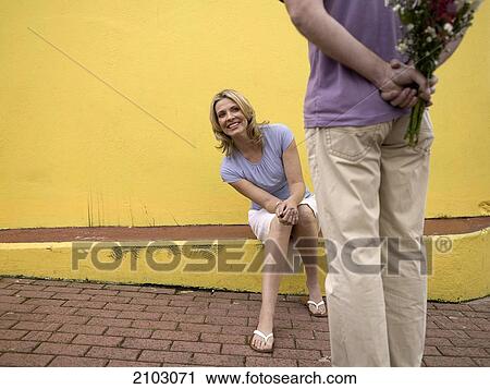 Stock Image - Woman looking at man hiding bouquet of flowers . Fotosearch