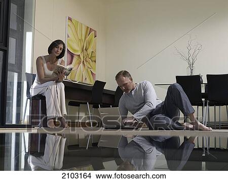 Picture - Woman reading book with her husband using laptop in living room . Fotosearch