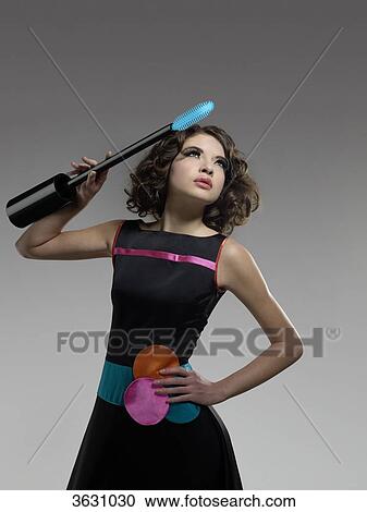 Stock Image - Young woman holding oversized mascara brush, composite . Fotosearch