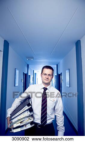Stock Image - Businessman standing in an office corridor, holding files, front view . Fotosearch