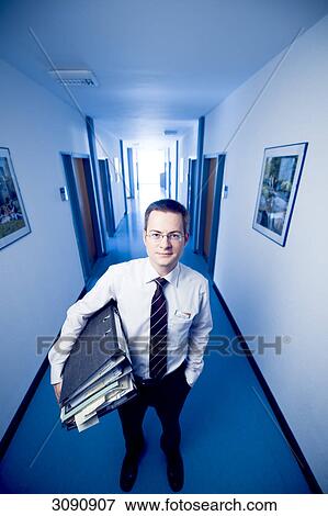 Businessman standing in an office corridor, holding files, front view View Large Photo Image Stock Photo - Businessman standing in an office corridor, holding files, front view . Fotosearch