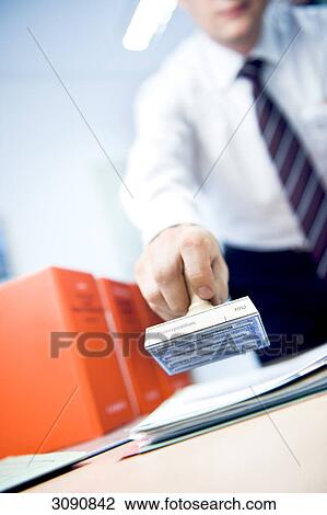 Stock Image - Man holding a stamp above a stack of papers, slanted view . Fotosearch