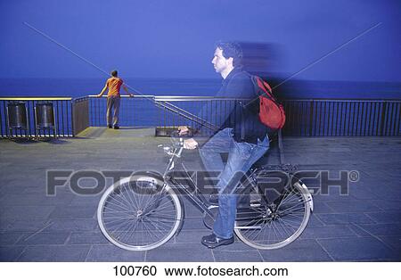 Stock Image - Side profile of man cycling on esplanade with another man standing in background, Barcelona, Spain . Fotosearch