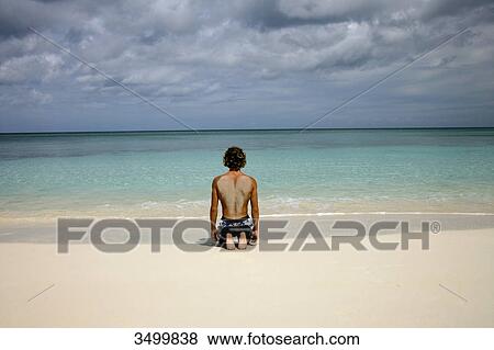Stock Photo - Man kneeling at the beach in front of ocean, Paradise Island, Bahamas, rear view . Fotosearch
