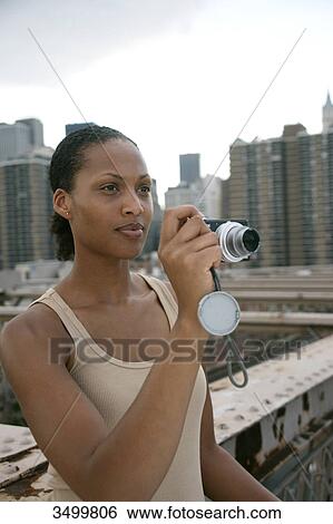 Stock Photograph - Woman standing on the Brooklyn Bridge and holding a digital camera, skyline of New York in the background, USA . Fotosearch