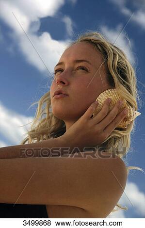 Stock Photo - Young woman holding a shell against her ear, low angle view . Fotosearch