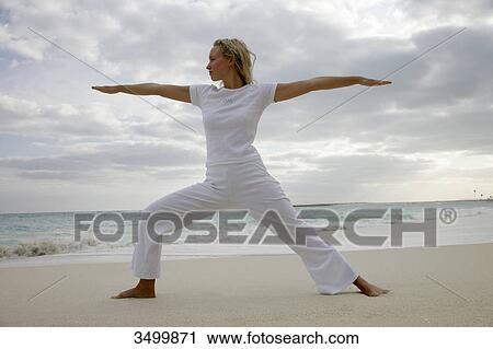 Stock Image - Young woman practising yoga on a beach, low angle view . Fotosearch
