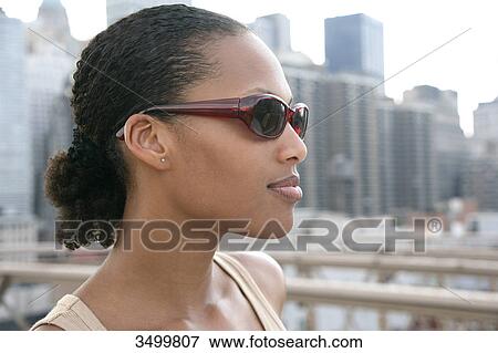 Stock Photo - Young woman wearing sunglasses, skyline in the background, portrait . Fotosearch
