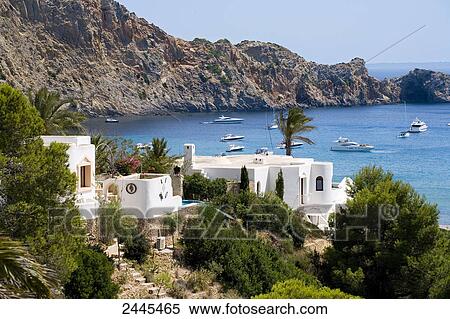 Stock Photography - Buildings at coast with sailboats in sea, Cala Jondal, Ibiza, Balearic Islands, Spain . Fotosearch