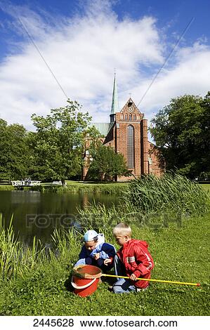 Stock Photo - Two children playing at pond side with church in background, Bad Doberan, Mecklenburg-West Pomerania, Germany . Fotosearch