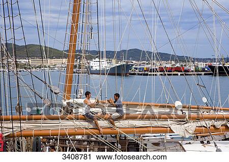 Stock Image - Two sailors sitting on a mast of a sailing ship, harbour in the background, Ibiza City, Ibiza, Spain . Fotosearch
