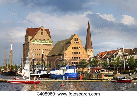 Stock Image - View of warehouses and St. Peter's Church, boats on a river in the foreground, Rostock, Germany . Fotosearch